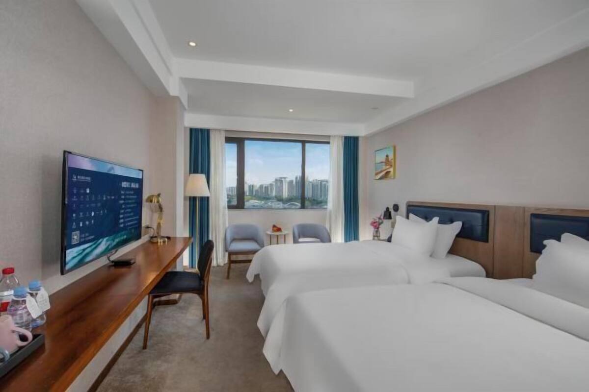 Habitación de hotel con un televisor grande, dos camas, un escritorio, una silla y ventana con vista a la ciudad.