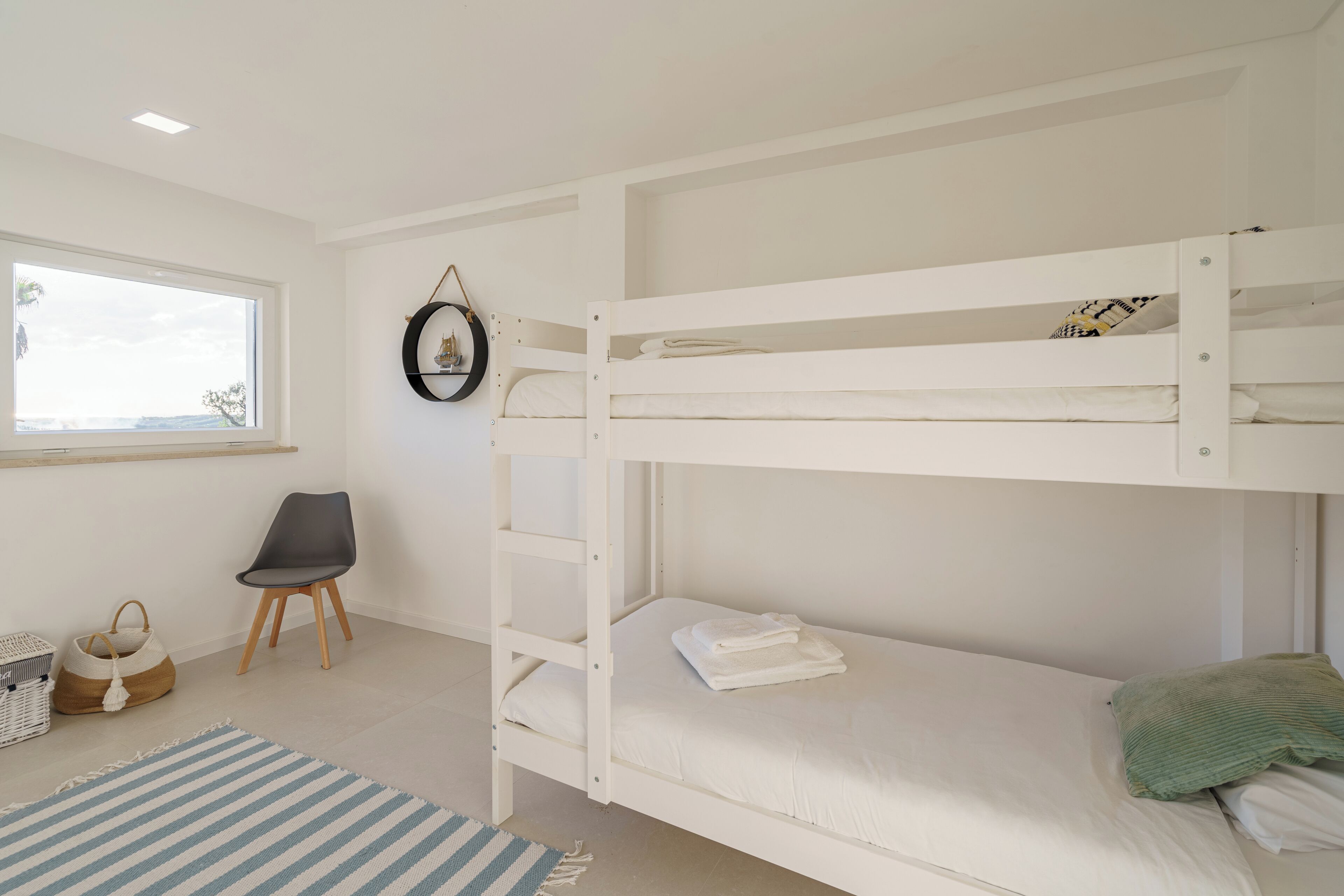 A bunk bed room with a window, a chair, a small rug, and a wall-mounted picture.