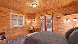 A wooden cabin room with a bed, a chair, a window with blinds, and a door leading outside.