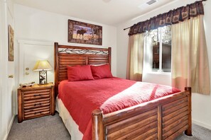 A wooden bed with a red blanket, a nightstand with a lamp, a framed picture on the wall, and a window with curtains.