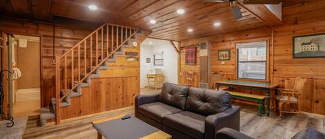 A wooden cabin interior with a staircase, a leather sofa, a wooden table, and a ceiling fan.