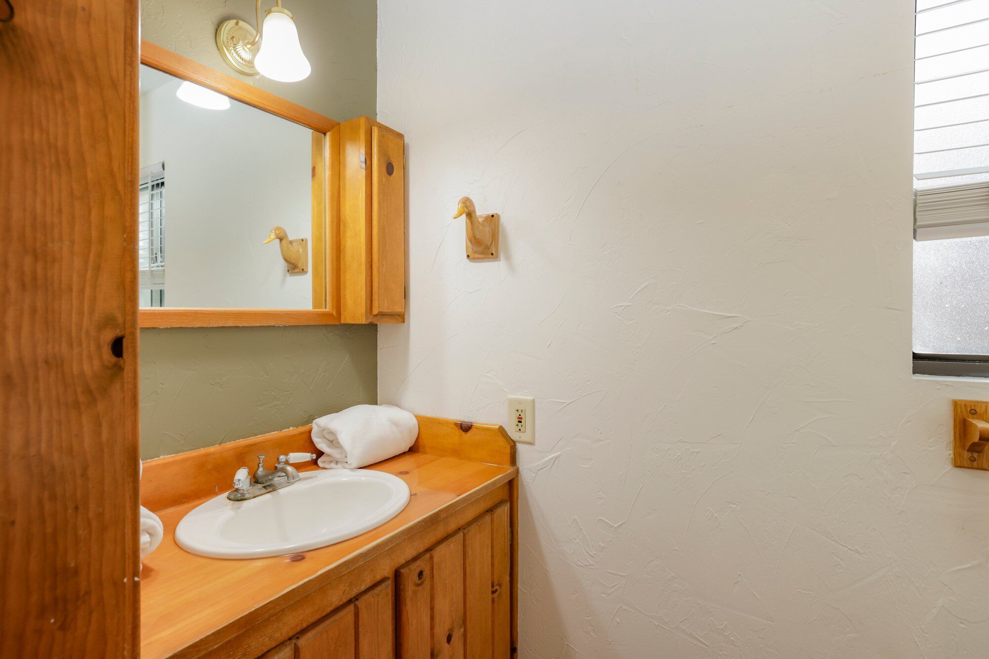 Un baño con tocador de madera, lavamanos blanco, espejo y lámpara sobre el espejo.