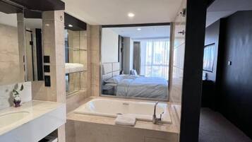 A modern bathroom with a large mirror, a bathtub, and a visible bedroom with a bed and curtains.