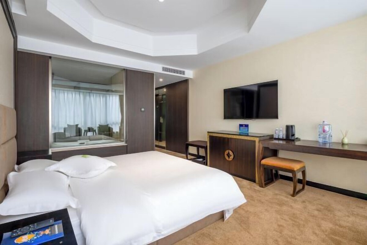 A hotel room with a large bed, a desk, a TV, and a view of the city through a large window.
