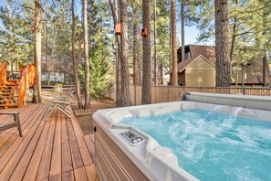 Een hottub op een houten terras, omgeven door hoge bomen, met een houten constructie en een gebouw op de achtergrond.