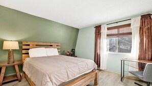 A bedroom with a wooden bed, a bedside table with a lamp, a desk with a chair, and a window with curtains.