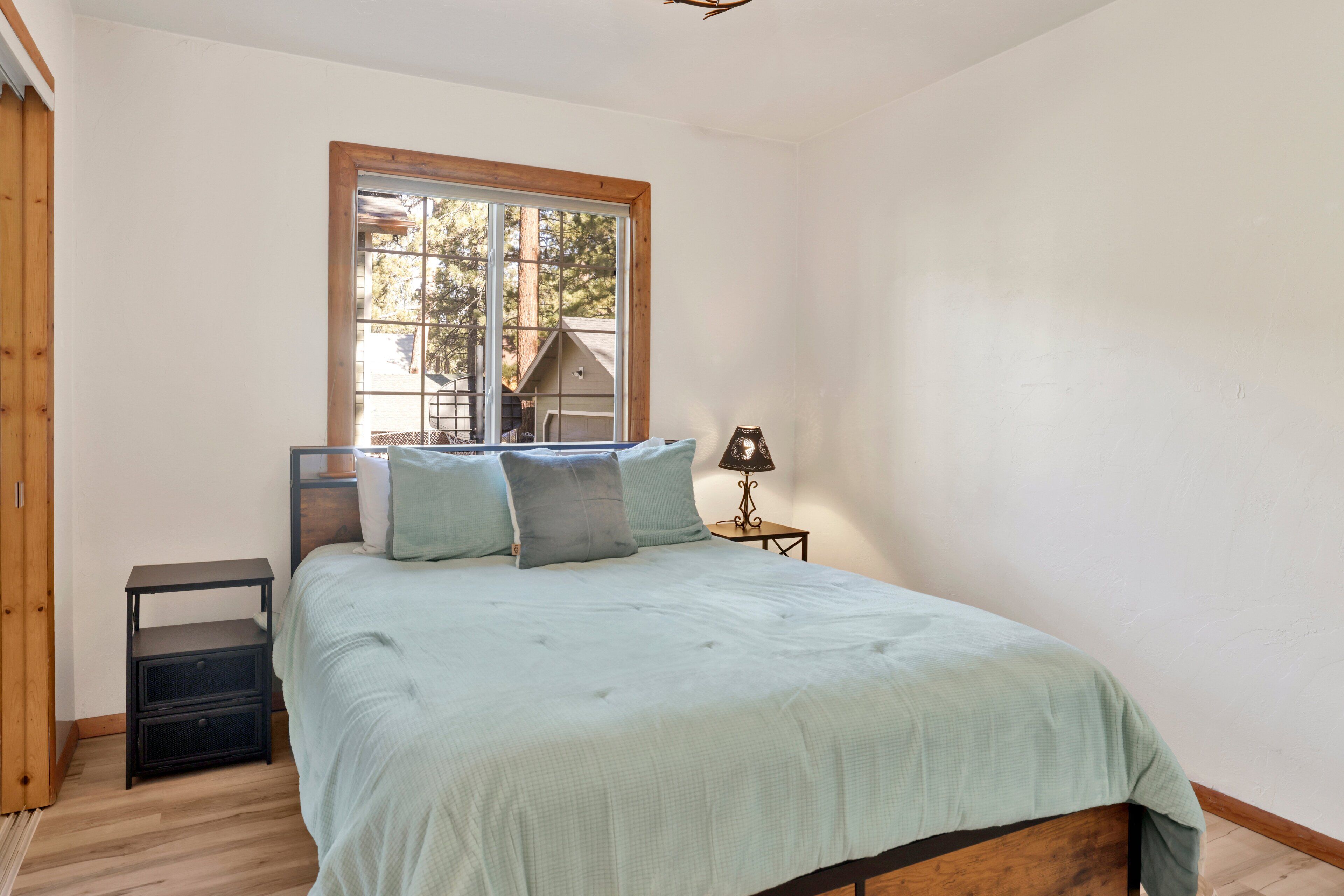 Une chambre à coucher avec un lit, une table de chevet, une lampe et une fenêtre donnant sur des arbres et une maison.