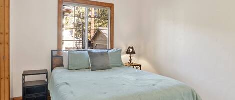 Une chambre à coucher avec un lit, une table de chevet, une lampe et une fenêtre donnant sur des arbres et une maison.