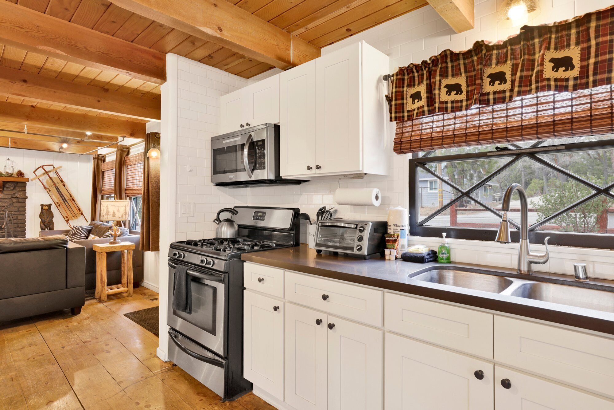 A modern kitchen with white cabinets, a stainless steel stove, a microwave, and a toaster. There is a window with a plaid curtain and a view of the outside.