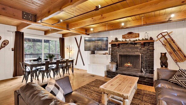 A cozy living room with a stone fireplace, leather sofa, wooden coffee table, and a dining area with a large TV. - Camp Conklin #2337 (Big Bear Lake)