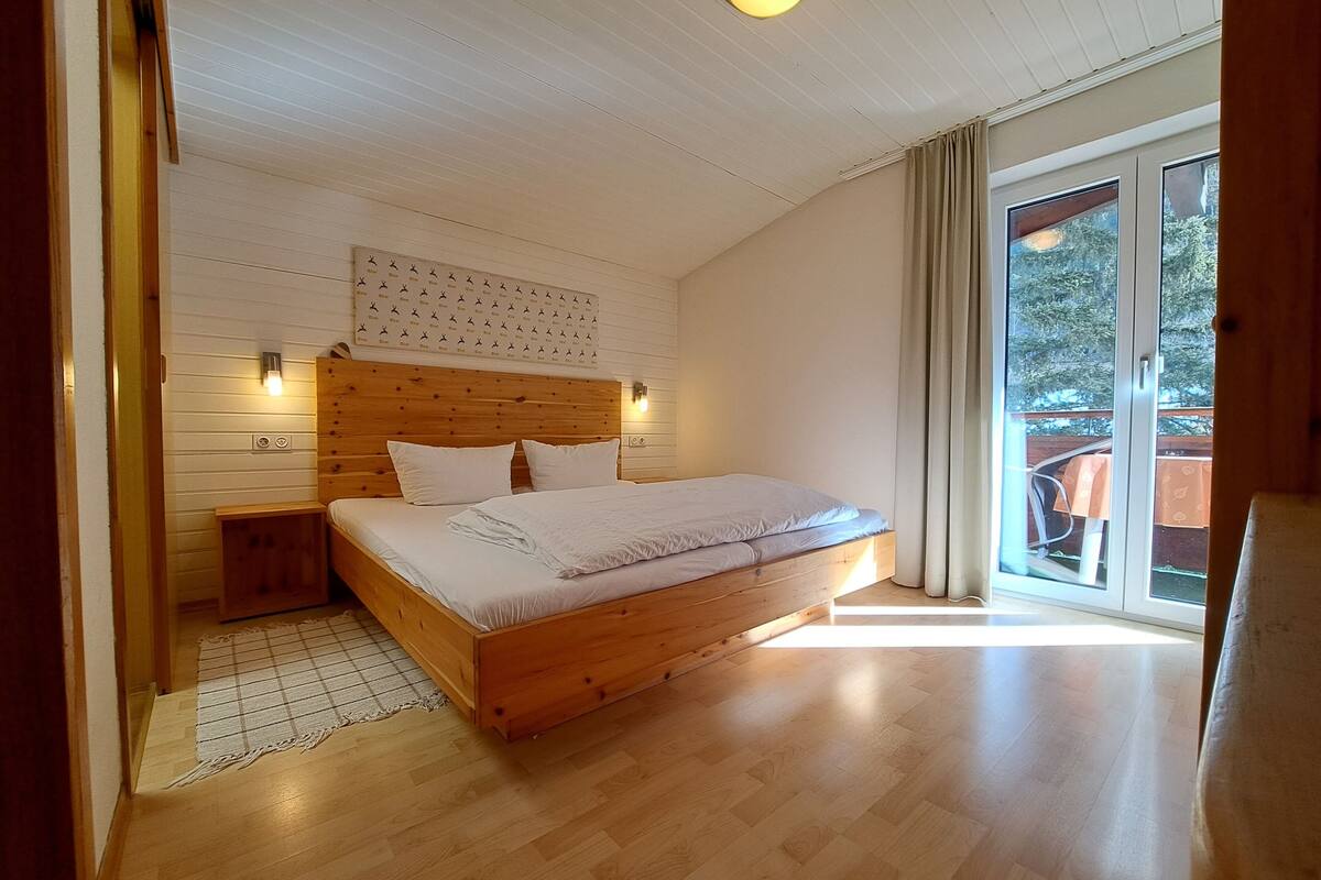 A bedroom with a wooden bed, a nightstand, a wall-mounted light, and a large window with curtains.