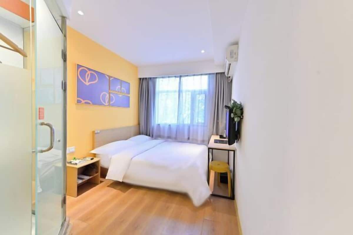 A hotel room with a bed, bedside table, a small stool, a desk with a chair, a window with curtains, and a wall with heart-shaped decorations.