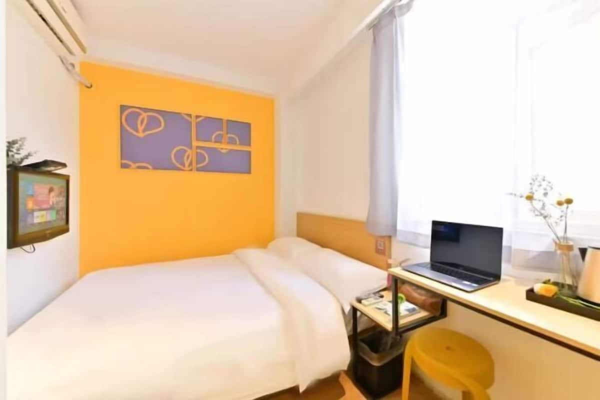 A hotel room with a bed, a desk with a laptop, a small trash bin, and a picture frame on the wall.