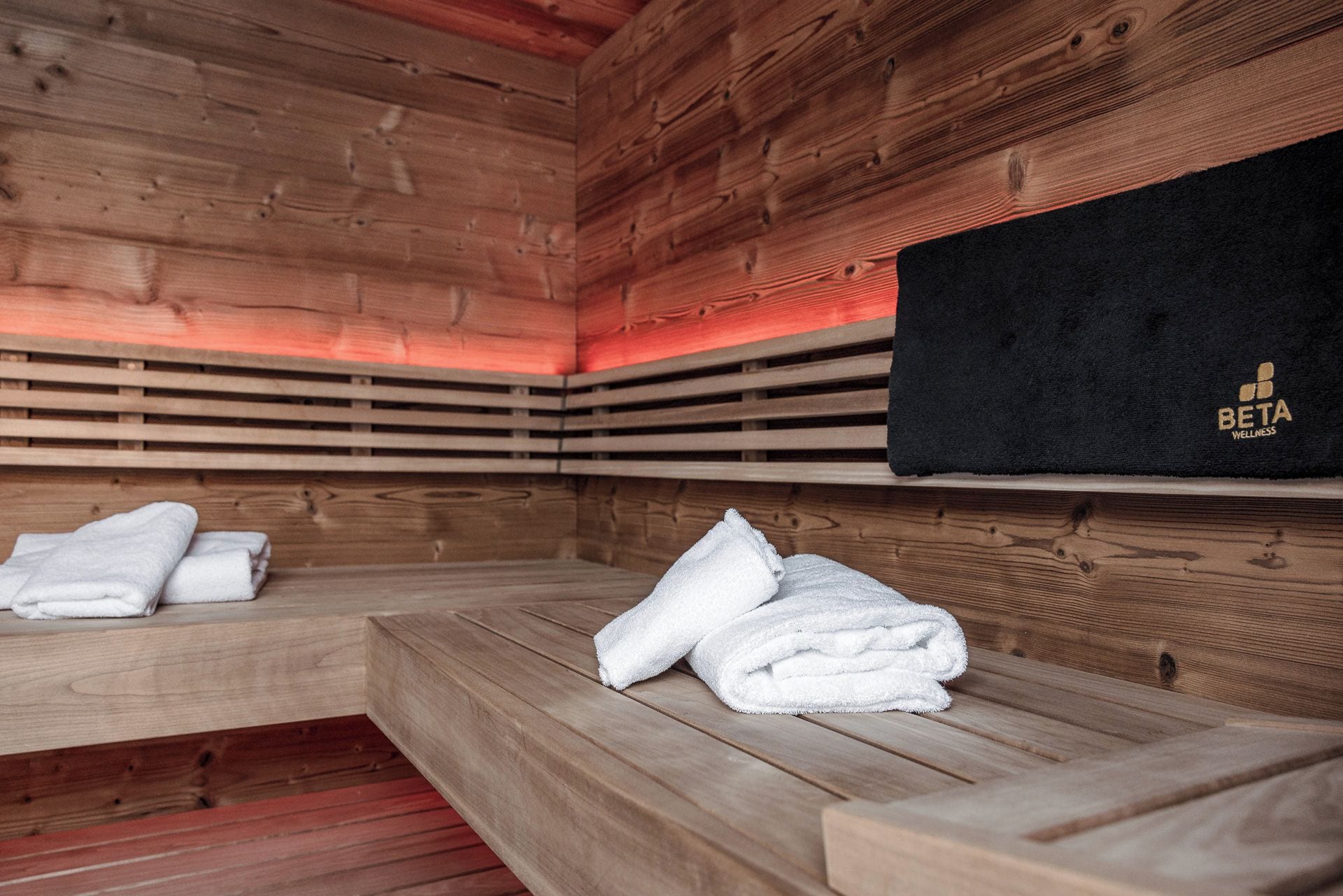 A sauna room with wooden benches and benches, a black towel rack with a logo, and a red light strip.