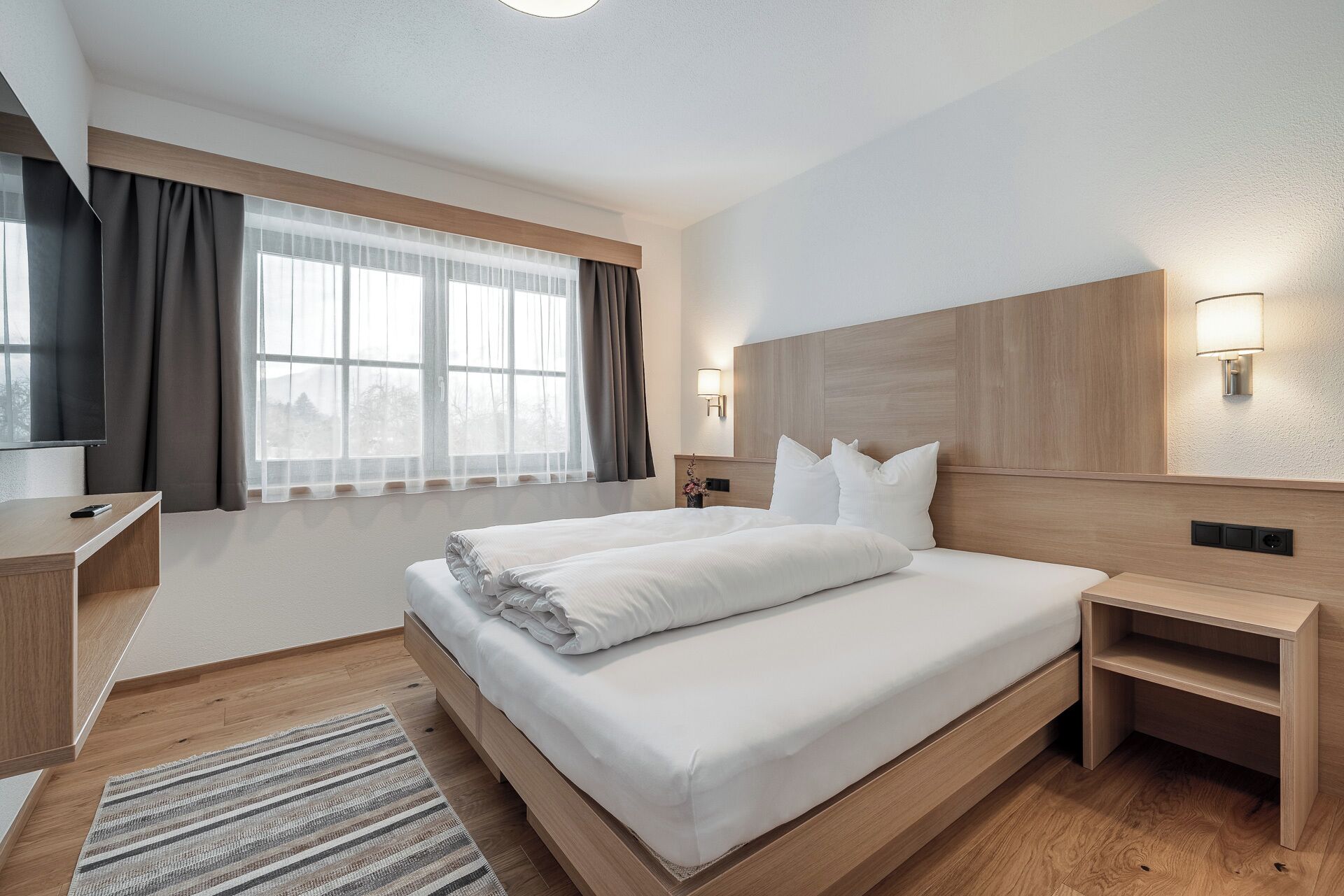 Ein modernes Hotelzimmer mit einem großen Bett, Nachttischen, einem Fernseher und einem Fenster mit Vorhängen.
