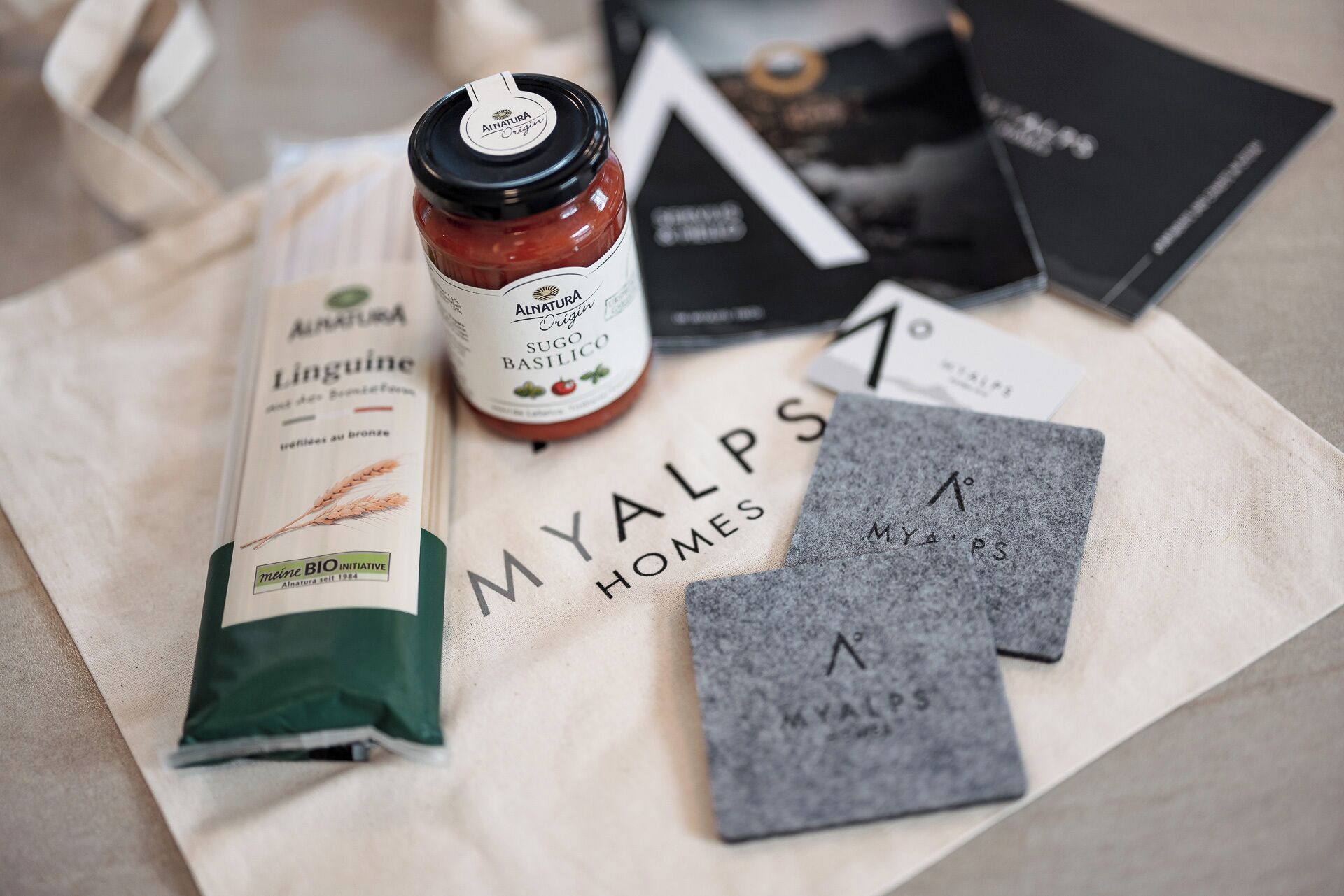 A table with a MyAlps Homes tote bag, a Linguiine cereal box, a Sugo Basilico jar, and a black booklet.