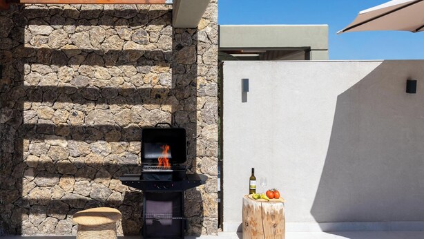 A stone wall, a barbecue grill, a wooden stool, and a round wooden table with food and drinks.
