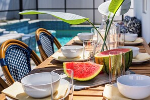 Une table à manger avec une pastèque, des bols, des verres et une bouteille, le tout disposé devant une vue sur la piscine.