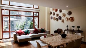 Un salon spacieux doté d’une grande fenêtre, d’un canapé agrémenté de coussins colorés, d’une table à manger et d’un mur décoré de chapeaux.