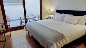 A hotel room with a large bed, a desk, and a chair. There is a view of the outside through a glass door.