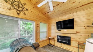 A cozy cabin interior with wooden walls, a ceiling fan, a TV mounted above a fireplace, and a large window offering a view of greenery.