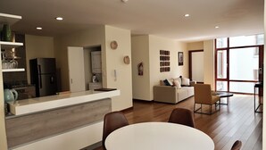 Eine moderne Küche mit integriertem Kühlschrank, ein Essbereich mit rundem Tisch und Stühlen, ein Wohnzimmer mit Sofa und Couchtisch sowie große Fenster, die für natürliches Licht sorgen.