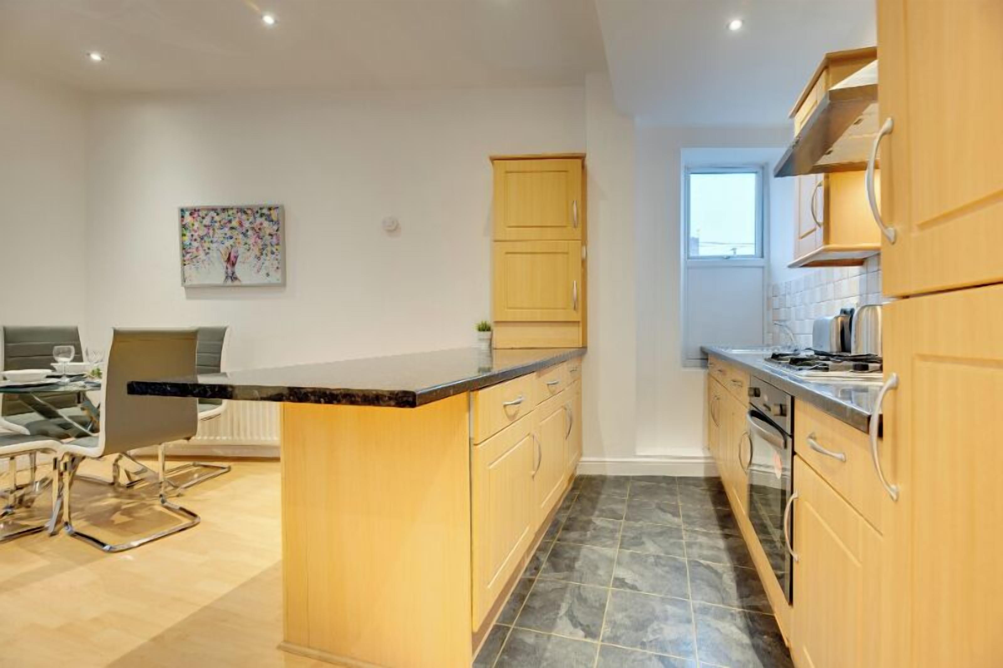 A modern kitchen with wooden cabinets, a marble countertop, and a dining area with a glass table and chairs.