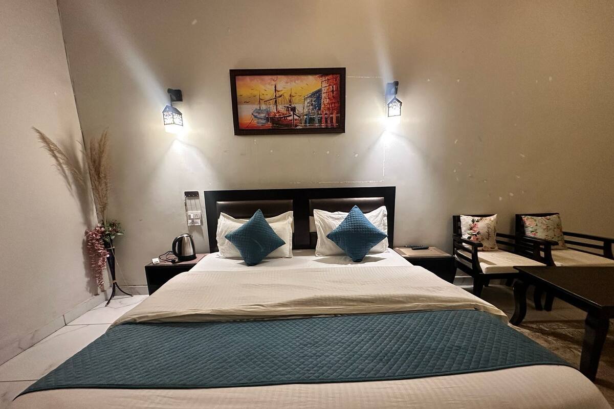 A neatly made bed with a blue quilt, two pillows, a bedside table with a lamp, a painting on the wall, and a chair with a floral cushion.