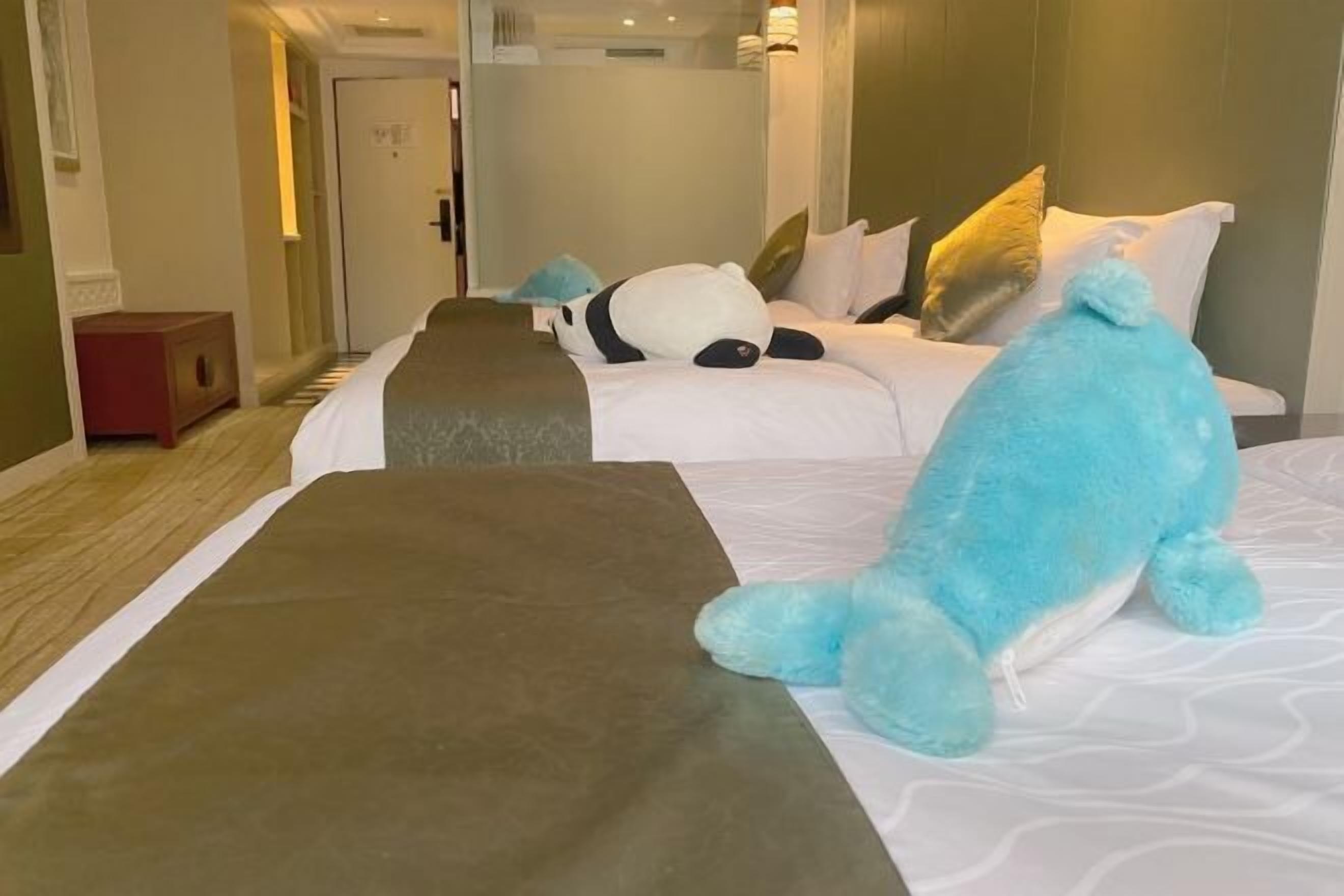A hotel room with two beds, each with a plush toy, a bedside table, and a door leading to another room.