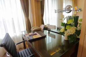 Ett hotellrum med ett skrivbord i glas, en stol, en telefon, ett anteckningsblock, en lampa och en vas med blommor.