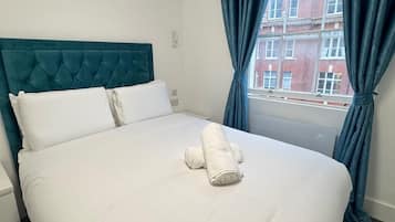 Habitación de hotel con una cama grande, cabecera color turquesa, ropa de cama blanca y vistas a edificios a través de una ventana con cortinas azules.