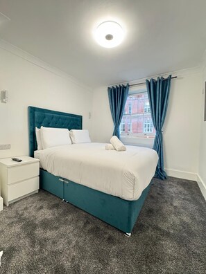 A bedroom with a teal upholstered bed, white bedding, a white bedside table, and a grey carpet. - Baker Street Apartments (London)