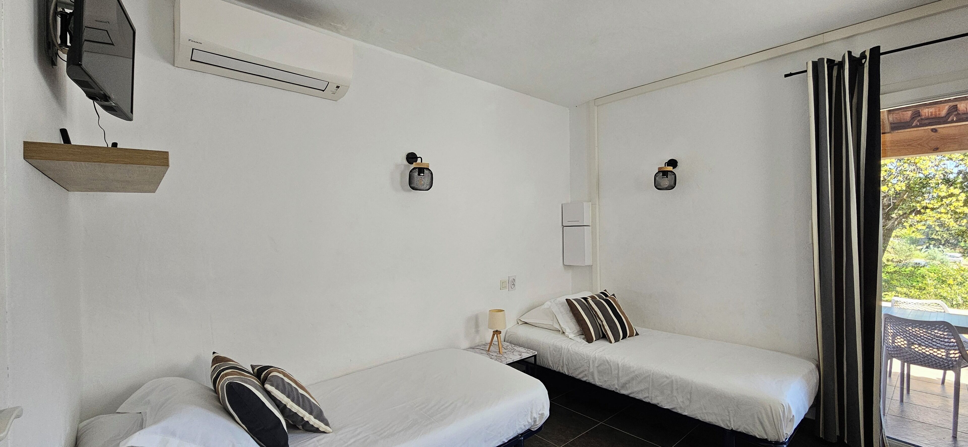 A room with two single beds, a mounted TV, an air conditioning unit, a small table with a lamp, and a view of greenery through a window.