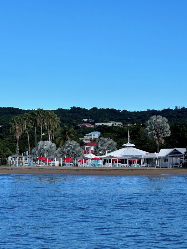 Un hotel frente a la playa con varios edificios blancos, sombrillas rojas y palmeras.