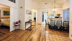 A spacious living area with a wooden floor, a dining table with chairs, a kitchen in the background, and a bed with a headboard.