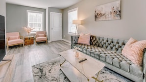 A living room with a grey tufted sofa, two pink armchairs, a coffee table, and a painting on the wall. - Charming Voyage: Ypsi & AnnArbor (Ypsilanti)