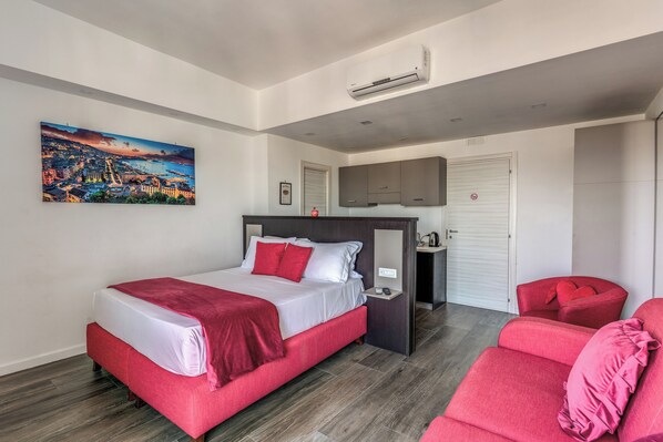 Une chambre d’hôtel moderne avec un grand lit, un canapé rouge, une petite table et un tableau représentant un paysage urbain accroché au mur.