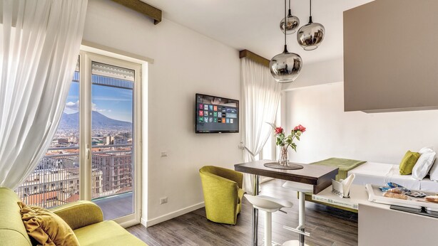 A modern living room with a green sofa, a dining area with a table and chairs, a flat-screen TV mounted on the wall, and a large window offering a city view. - Timeless (Naples)