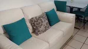 A living room with a beige sofa, teal cushions, and a patterned throw pillow. There is a small table with chairs and a potted plant. - Small Independent Studio in Zapopan (Zapopan)