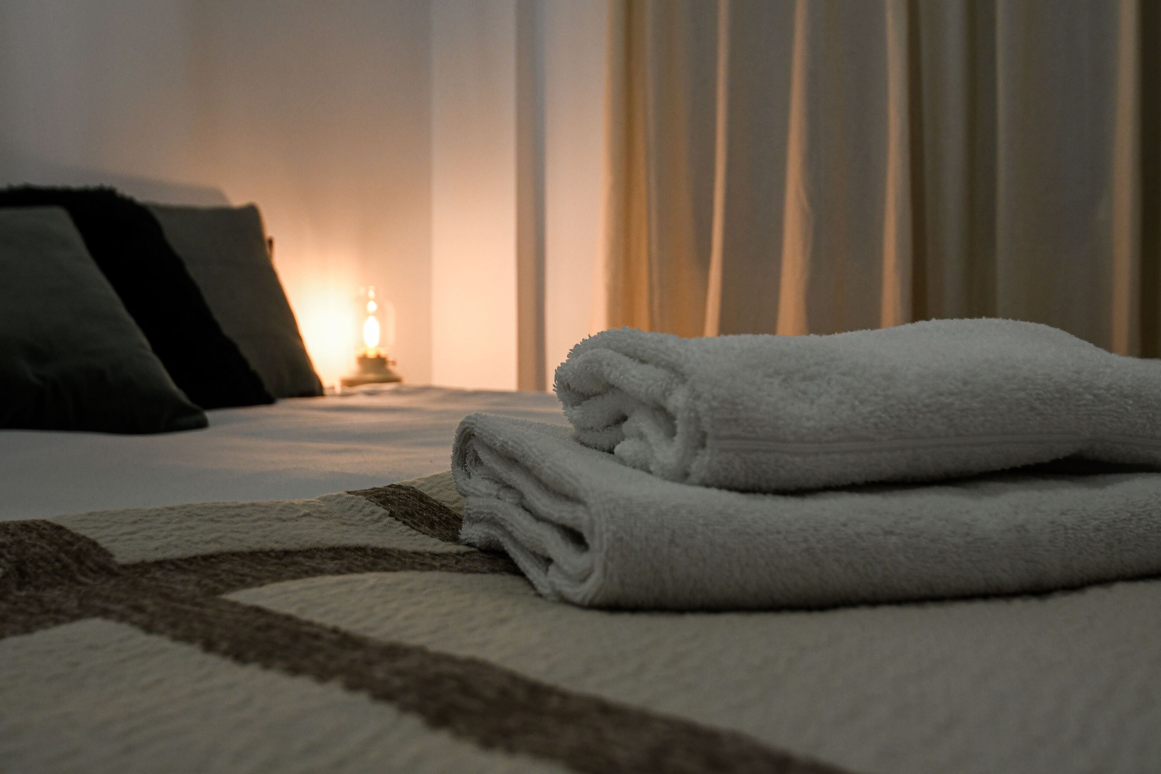 Una cama bien tendida con una toalla blanca doblada, una lámpara de noche y una ventana con cortinas.