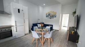 A modern kitchen with white appliances, a dining table with chairs, a blue sofa, and a flat-screen TV.
