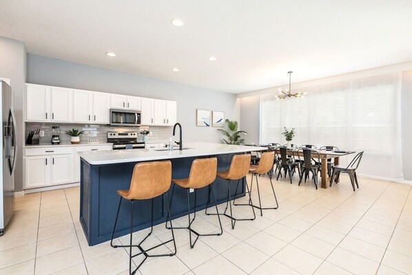 A modern kitchen with white cabinets, a central island with bar stools, a dining area with a wooden table and chairs, and a large window with blinds. - Family 6BR Resort Villa | Pool & Spa near Disney (Kissimmee)