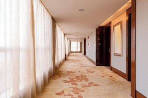 A hotel corridor with a carpeted floor, large windows with curtains, and doors on the right.