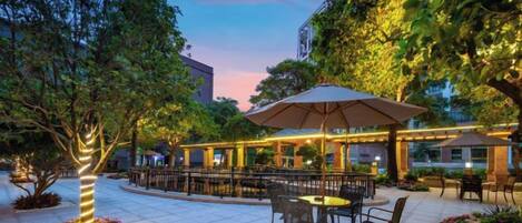 An outdoor patio area with tables and chairs, surrounded by trees and a covered seating area.