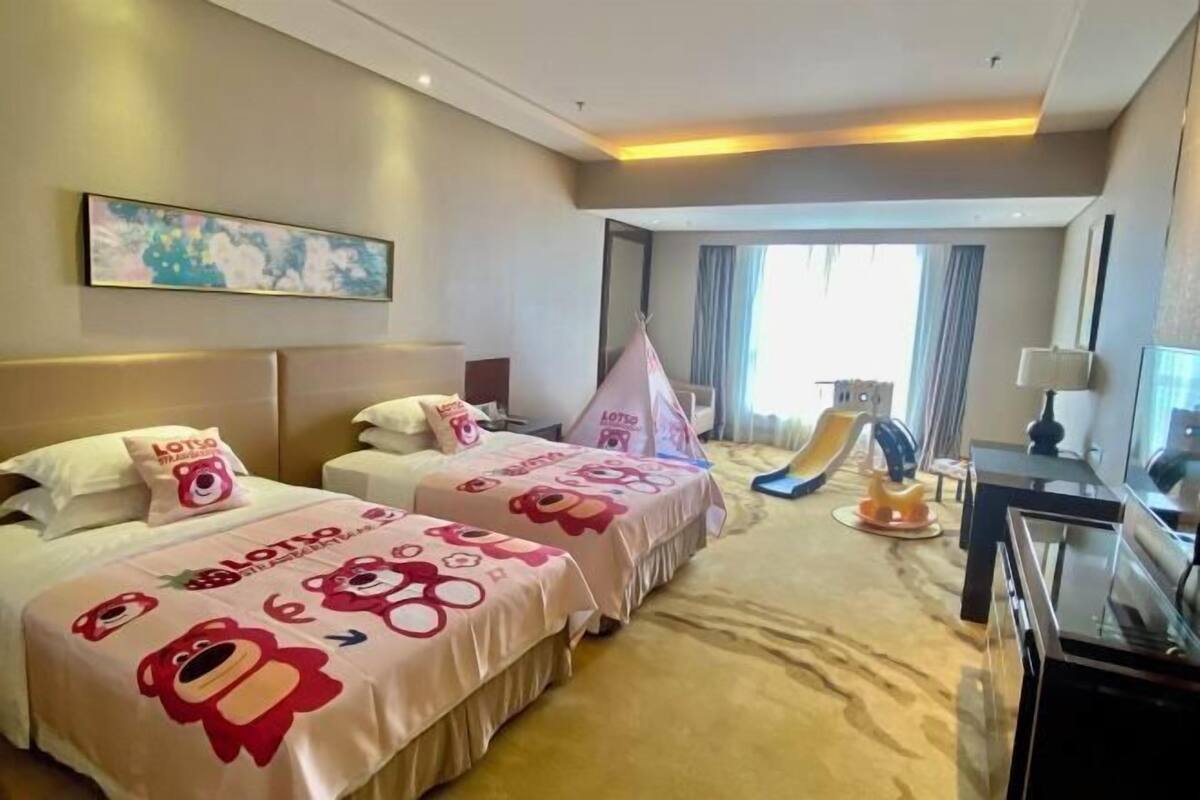 A hotel room with two beds, a desk, a chair, a desk lamp, a window with curtains, and a painting on the wall.