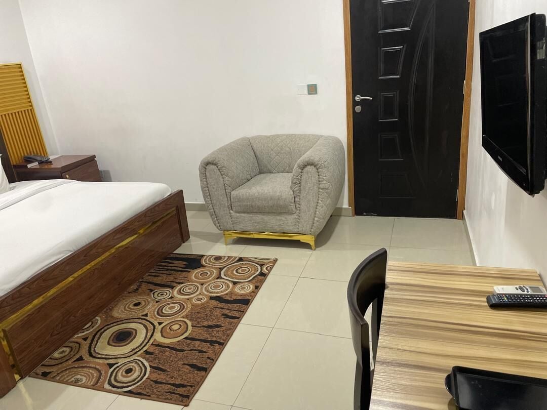 A hotel room with a bed, a grey armchair, a wooden desk, and a TV.