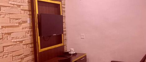 Una stanza modesta con una parete a effetto pietra, una scrivania in legno con una sedia, un televisore a parete, un divano rosso e un condizionatore d'aria.