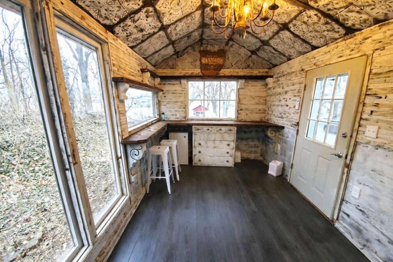 A rustic cabin interior with a stone ceiling, wooden walls, a bar area with stools, and a window overlooking a forested area.
