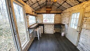 A rustic cabin interior with a stone ceiling, wooden walls, a bar area with stools, and a window overlooking a forested area.