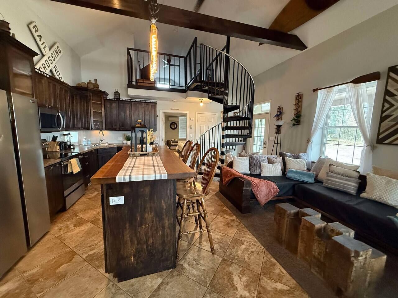 A spacious kitchen with a central island, a dining area with chairs, a staircase, and a living area with a sofa and cushions.